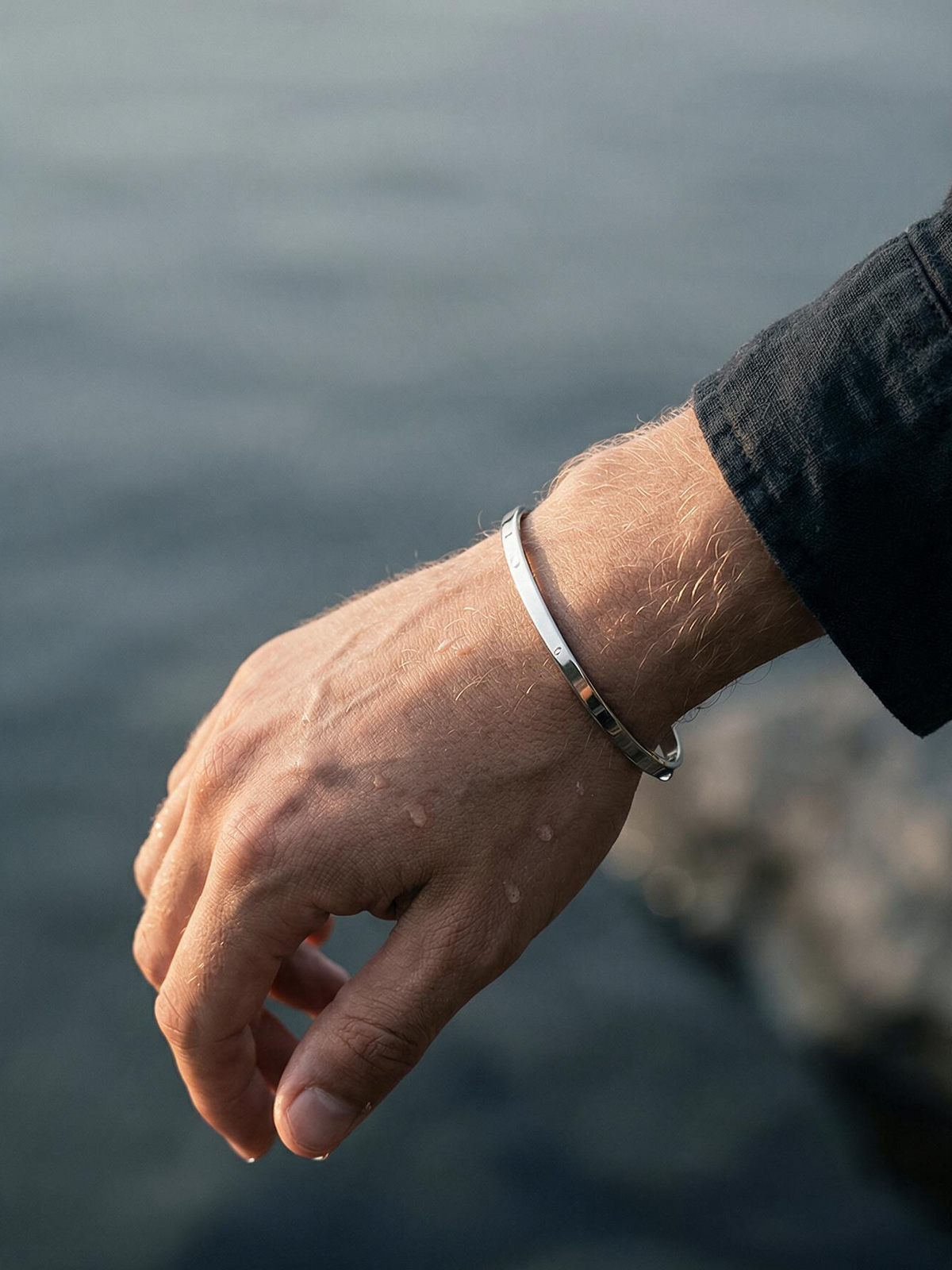Brazalete plateado en la muñeca junto al mar, acero quirúrgico resistente al agua.