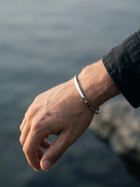 Brazalete plateado en la muñeca junto al mar, acero quirúrgico resistente al agua.