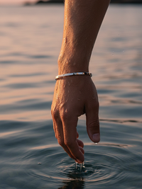 Brazalete plateado en la muñeca tocando el agua al atardecer, resistente y duradero.